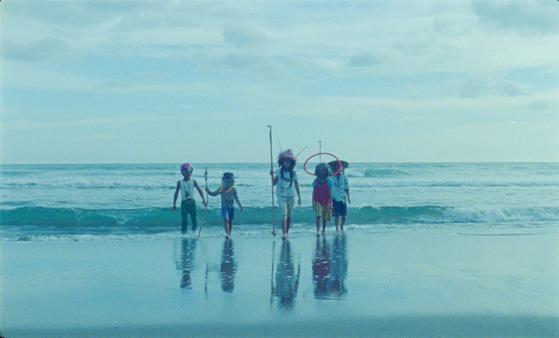A row of 5 children standing by the waves wearing different ornamental headgear