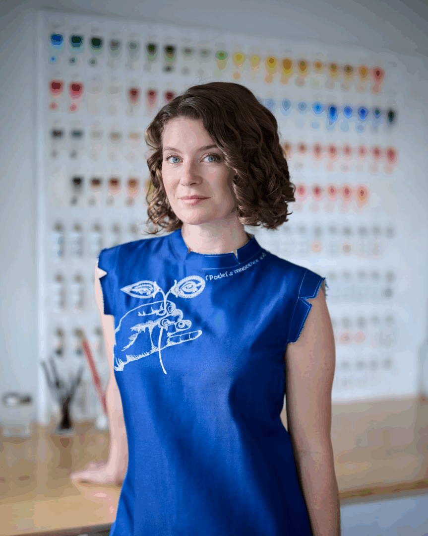White female with brown hair wearing a blue shirt stands before a blurred color chart.