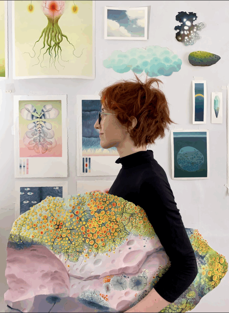 White woman with red hair stands in profile holding a painting and stands before a wall with art on it.